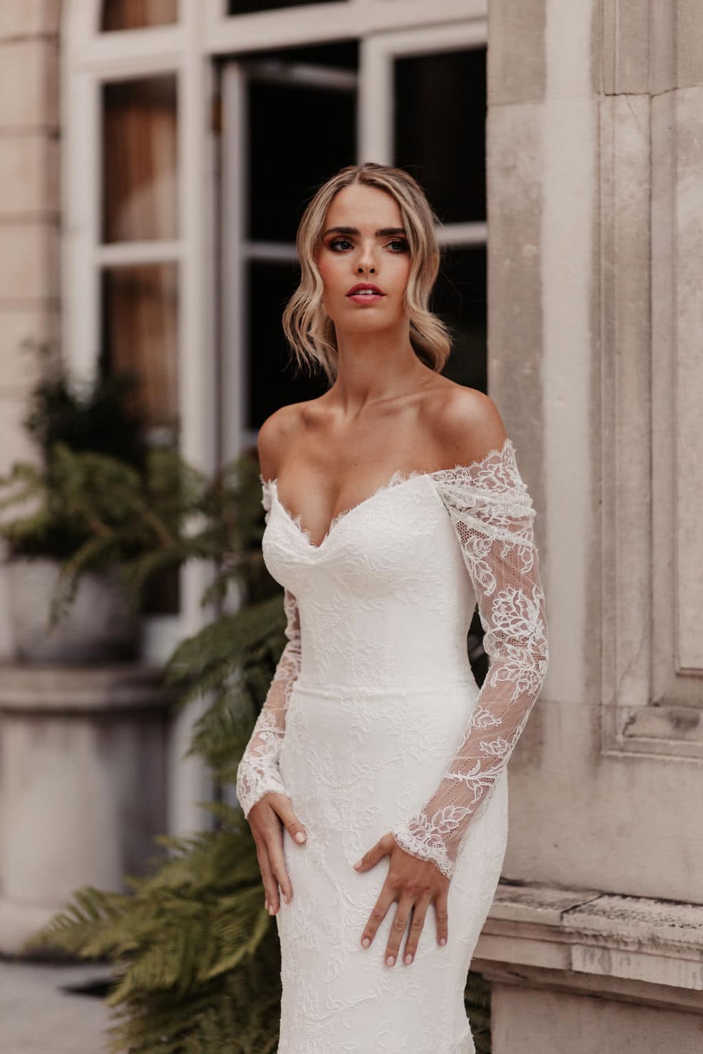 Showstopping Suzanne Neville Stravinsky has a gorgeous scoop off the shoulder neckline with a corseted cupped bodice, detachable long lace sleeves, a fitted lace skirt. Find your dream wedding dress at Your Dream Bridal near Boston.